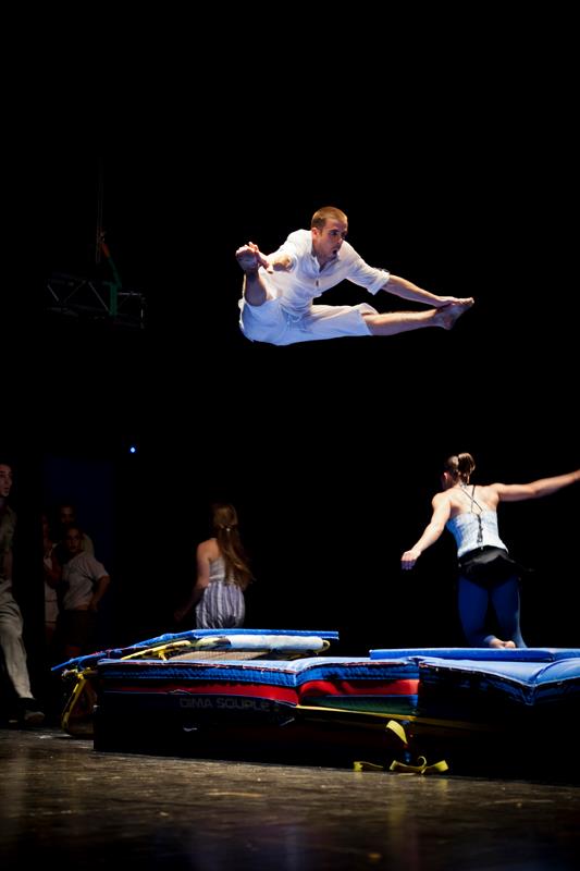 Mikelo, the multidisciplinary circus artist, performs an impressive acrobatic jump in mid-air above safety mats on stage.