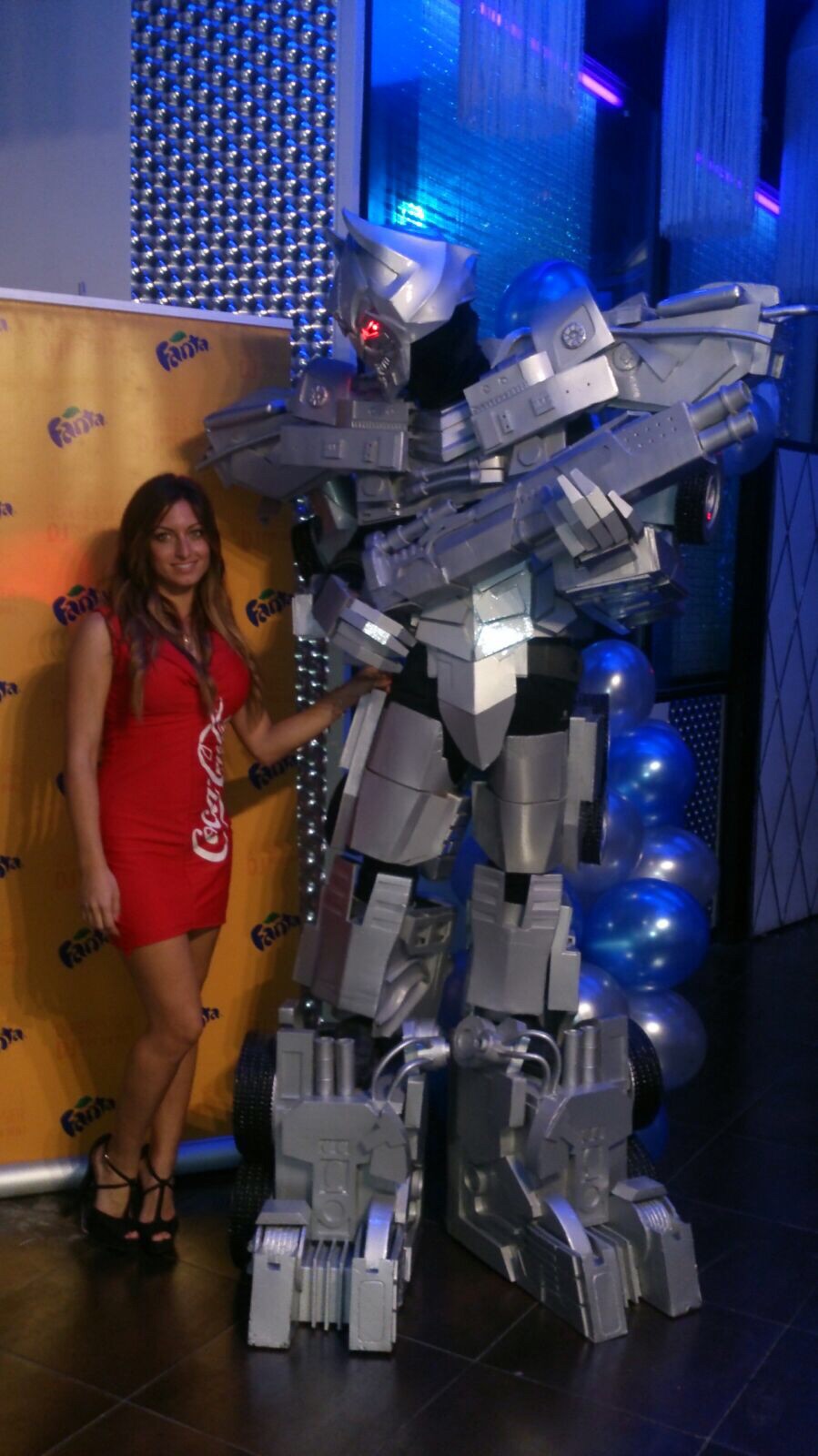 Circus artist Mikelo's silver robot performer with glowing red eyes poses with a woman in a red Coca-Cola dress at a branded 