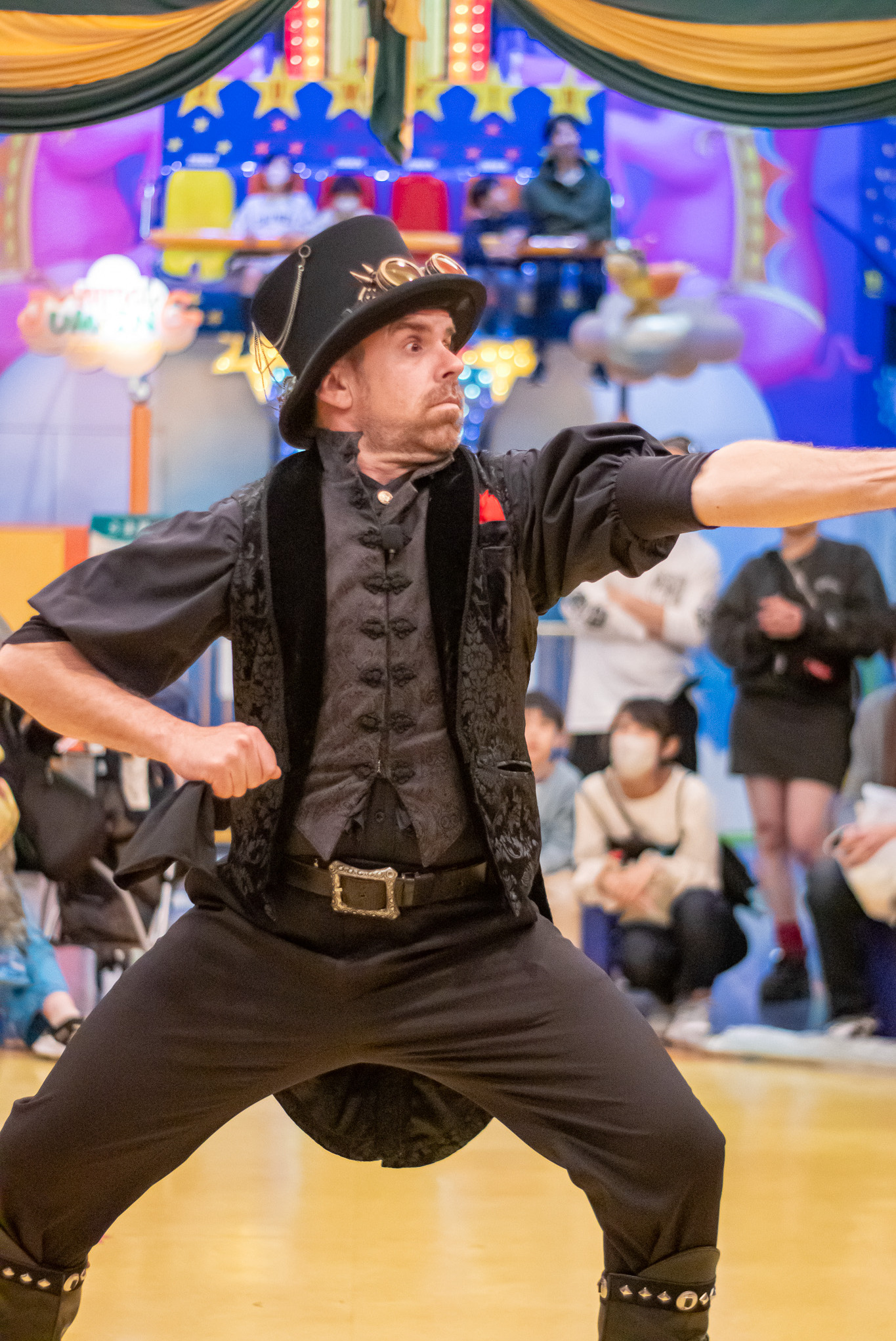 Mikelo, a circus artist, performs in a black steampunk costume, striking a dynamic pose on stage with an intense gaze.