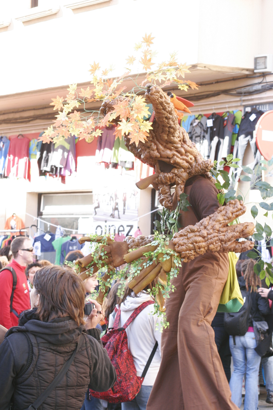 Mikelo, a circus artist, performs on stilts in a detailed tree costume with autumn leaves and vines for a street audience.