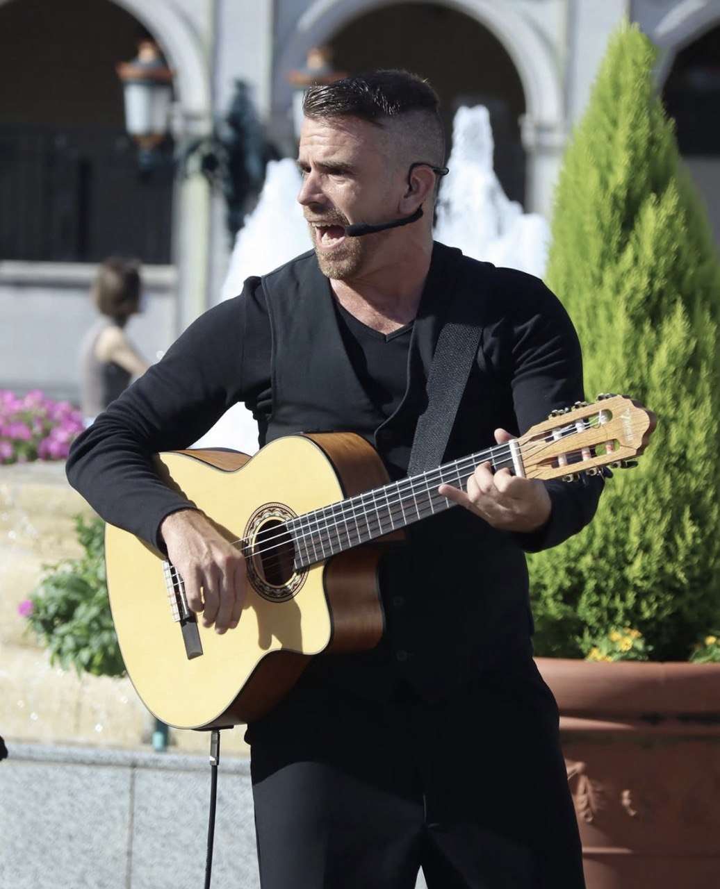 Multidisciplinary artist Mikelo performs live, singing into a headset mic while playing an acoustic guitar in a black vest.